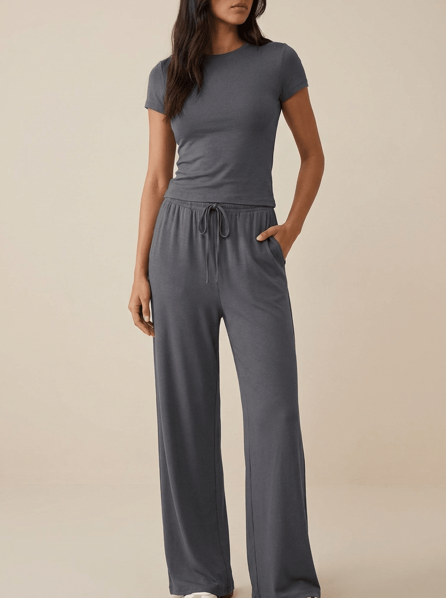 A woman modeling a dark gray short-sleeve crewneck top and matching wide-leg drawstring pants against a solid beige studio background.